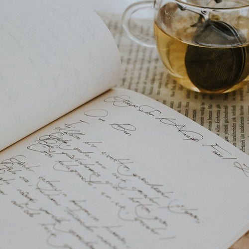 An open journal beside loose herbal tea leaves and a glass cup on vintage paper